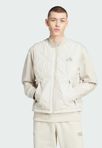 Chaqueta bomber acolchada en color beige claro con puños acanalados, dos bolsillos frontales y cierre de cremallera. Presenta un sutil logo en el pecho.