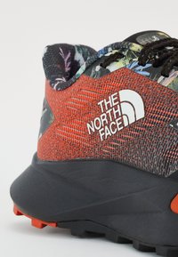 La chaussure de trail présente un dessus texturé rouge avec des accents floraux, une semelle noire et un logo blanc. Un design léger avec une adhérence robuste.