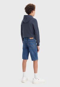 Marinblå kort hoodie, denimshorts, vita sneakers. Texturerad tyg, synliga sömmar och märkesetikett på shortsen. Avslappnad klädstil, bakvy.