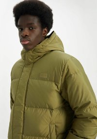 Chaqueta acolchada verde oliva con cuello alto, cierre de cremallera, patrón acolchado y un pequeño logotipo en el pecho. Textura suave y lisa.