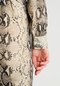 Beżowa i czarna bluzka w wężowy wzór z długimi rękawami, teksturowaną tkaniną i dopasowanymi mankietami, ukazująca dłoń na dole.