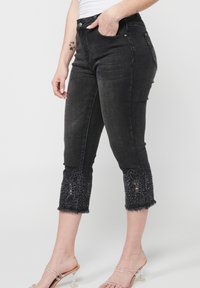 Crne traperice capri dugih nogavica imaju vezene čipkaste detalje na rubovima, rese i uski kroj s džepovima.