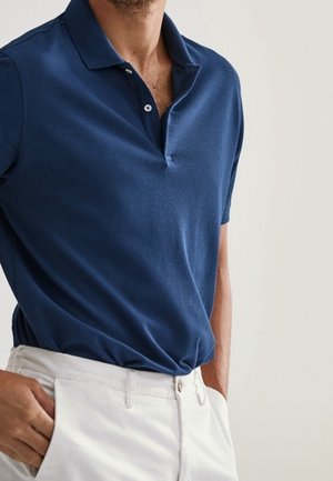 Mies, jolla on päällään laivastonsininen, lyhythihainen poolopaita, joka on laitettu valkoisten housujen sisään, kädet taskuissa, yksinkertaisen taustan edessä.