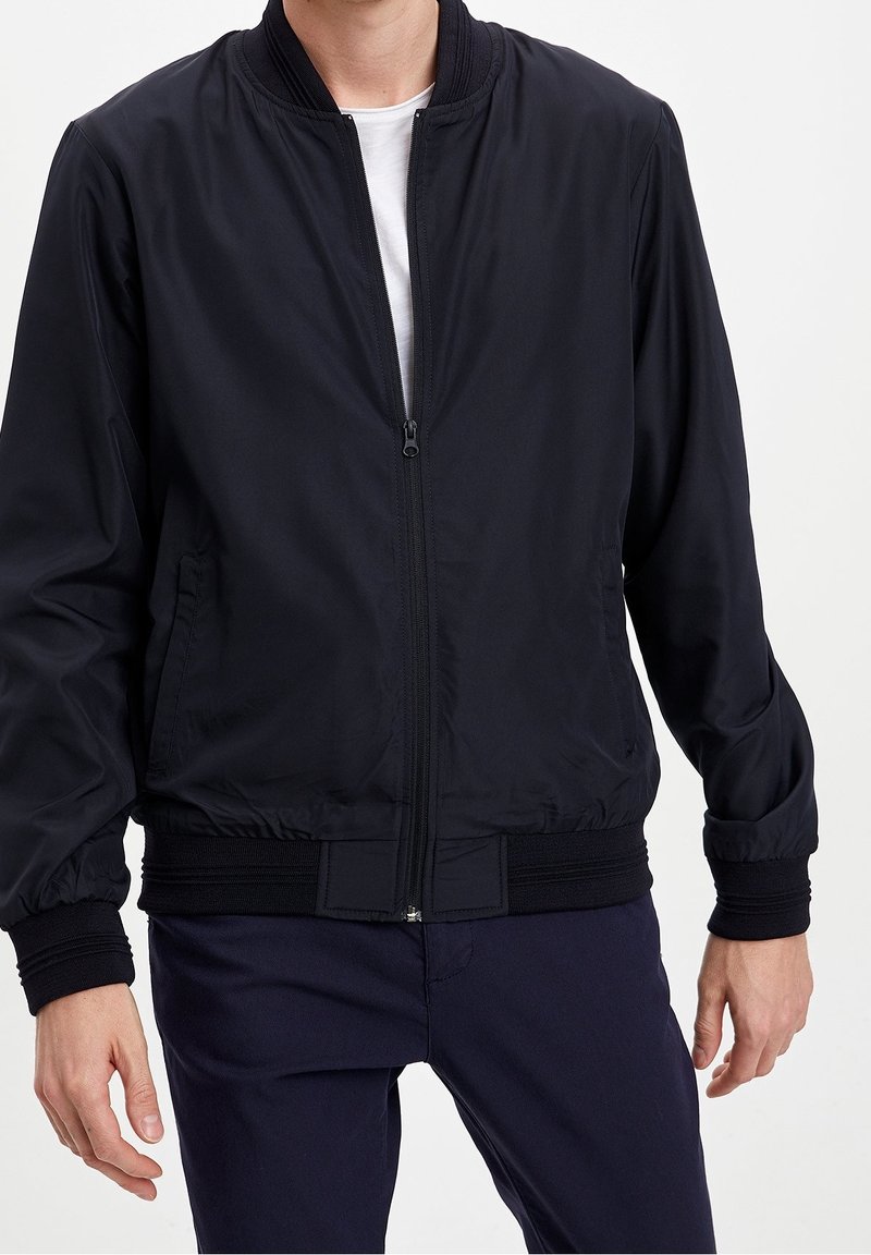 Homme portant une veste bomber noire zippée avec poignets et ourlet côtelés, par-dessus un t-shirt blanc et un pantalon sombre.