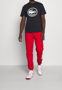 Schwarzes Baumwoll-T-Shirt mit weißem Lacoste-Logo, kombiniert mit leuchtend roten Jogginghosen mit Farbblock-Details. Weiße und rote Sneaker runden das Outfit ab.