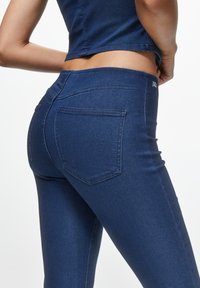 Kvinna klädd i mörkblå skinny jeans och en matchande kort topp, visad från midjan och vinklad åt sidan.