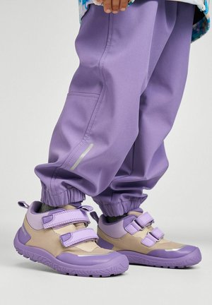 Dziecko noszące beżowo-fioletowe tenisówki na rzepy oraz fioletowe spodnie dresowe ze ściągaczami przy kostkach, stojące na jasnym tle.