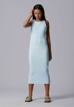 Joven mujer de pie, vestida con un vestido midi sin mangas de color azul claro, zapatos beige sin cordones, collar de oro y pendientes sobre un fondo gris.