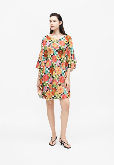 Γυναίκα στέκεται φορώντας ένα χαλαρό φόρεμα μέχρι τα γόνατα με πορτοκαλί, ροζ και πράσινα λουλούδια και μαύρα πέδιλα με τακούνι.