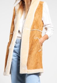 Tan-färgad fuskpälsväst med gräddfärgade detaljer, öppen framtill, två sidofickor och avslappnad passform. Strukturerad tyg med en mjuklook.