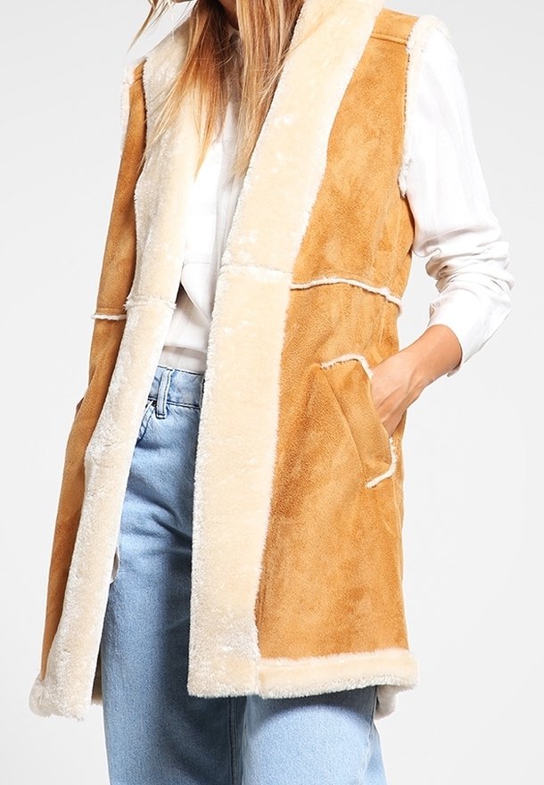 Tan-färgad fuskpälsväst med gräddfärgade detaljer, öppen framtill, två sidofickor och avslappnad passform. Strukturerad tyg med en mjuklook.