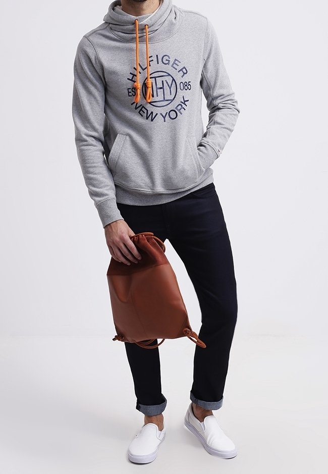 Homme portant un sweat à capuche gris Tommy Hilfiger, pantalon noir retroussé, chaussures blanches sans lacets, tenant un sac à cordon en cuir marron.