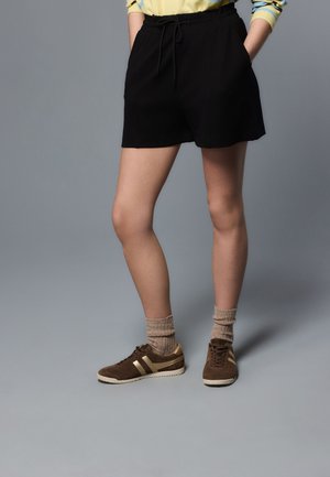 Personne portant un short noir, des baskets marron avec des rayures beige, des chaussettes beiges et une chemise jaune avec des rayures bleues sur les manches.