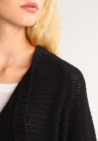 Blondýnka v černém pleteném kardiganu přes bílé tričko, zaměřená na rameno a dolní část obličeje na neutrálním pozadí.