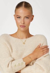 Goldene Halskette mit rundem Anhänger und passendem Armband. Das Modell trägt einen cremefarbenen Pullover mit grobem Strick und goldenen Knopf-Akzenten.