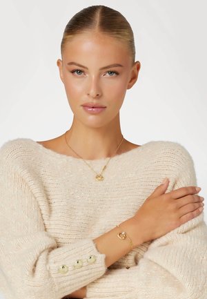 Collana d'oro con pendente circolare e bracciale coordinato. Il modello indossa un maglione crema con lavorazione a trecce e dettagli in oro.