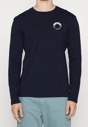 Osoba nosząca dopasowaną czarną koszulkę z długim rękawem z białym logo "Scotch & Soda" na lewej piersi oraz jasnoniebieskie spodnie.