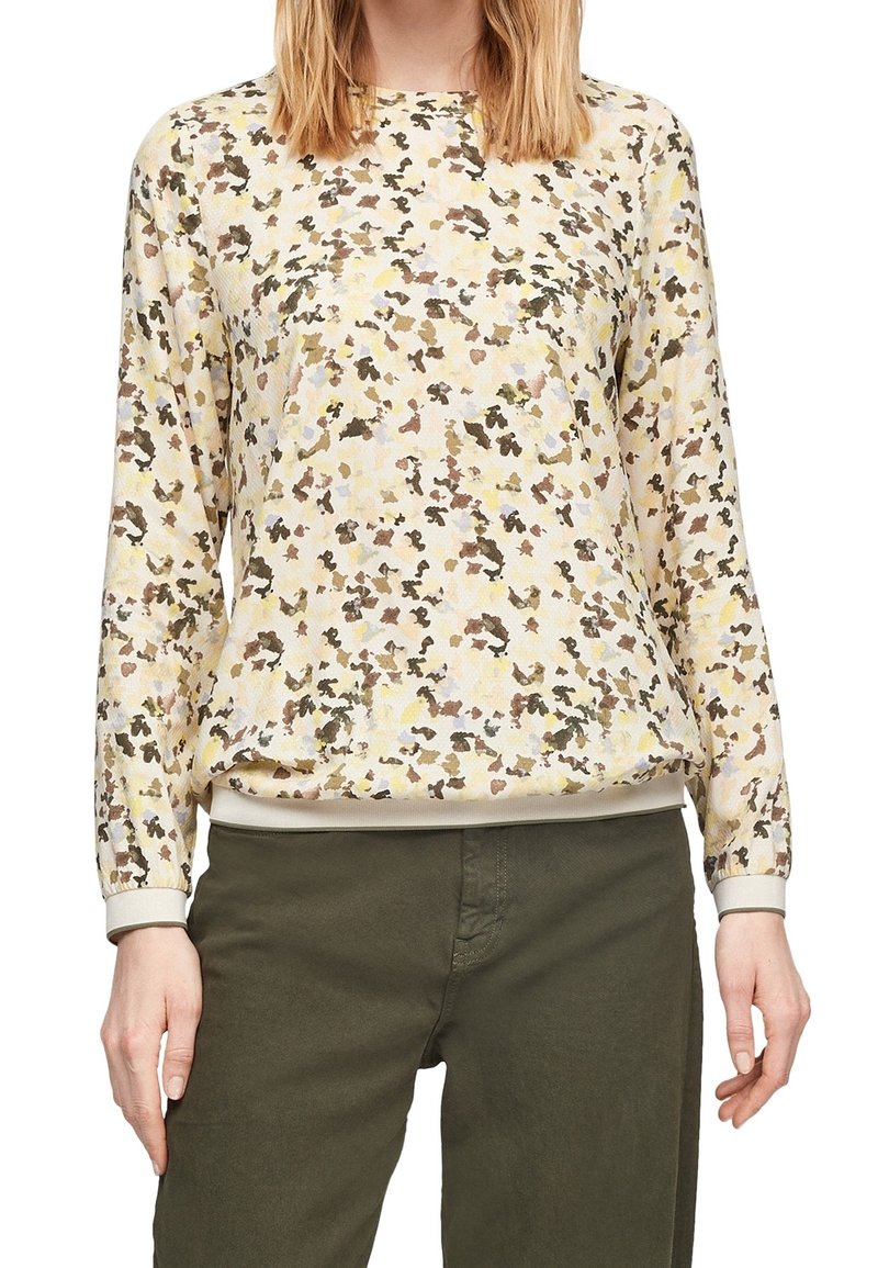 Femme portant un pull à manches longues beige et marron à motif floral et un pantalon vert olive, debout devant un fond blanc.