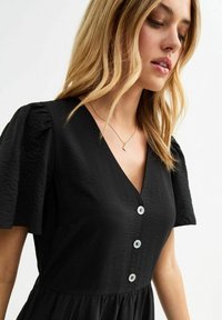 Vestido negro con escote en V, mangas cortas abullonadas y seis grandes botones de color claro en el frente. Tela texturizada con una silueta fluida.