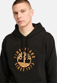 Hoodie in cotone nero con logo ricamato raffigurante un albero in oro. Cappuccio con cordino regolabile e polsini a costine.