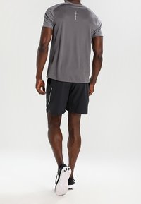 Graues sportliches T-Shirt mit einem kleinen vertikalen reflektierenden Detail, kombiniert mit schwarzen Shorts und Sportschuhen mit weißer Sohle und schwarzen Akzenten.