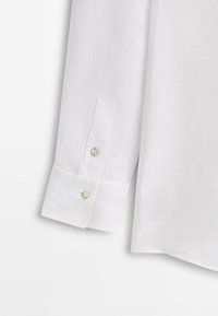 Cuff di una camicia bianca a maniche lunghe con due bottoni simili a perle su uno sfondo di tessuto semplice, che mostra dettagliata cucitura e texture.