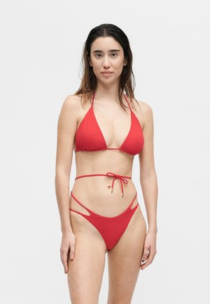 Žena s dlhými tmavými vlasmi, ktorá má na sebe červené tangá v tvare trojuholníka s výrezmi a vysokým pásom na jednoliatom svetlom pozadí.