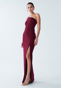 Bordo obleka brez naramnic, oprijeta ob telesu, s stranskim razporkom, iz gladkega blaga, dopolnjena z zlatimi sandali s tankimi trakovi in visokimi petami.