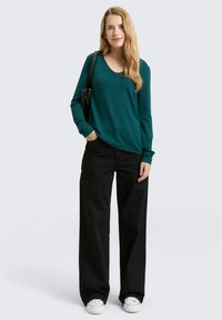 Turquoise trui met V-hals en lange mouwen, gecombineerd met zwarte, hooggetailleerde, brede broekspijpen en witte sneakers. Simpele zwarte handtas.