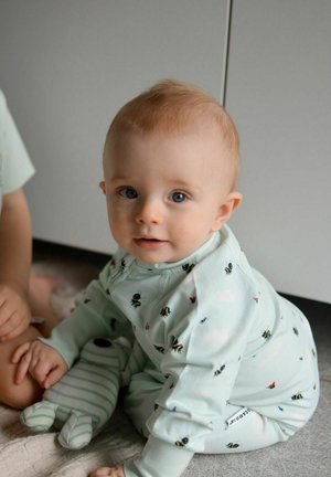 Vaaleanvihreät vauvan pyjamat, joissa on mehiläisten ja perhosten kuvioita, pehmeästä kankaasta ja pitkät hihat. Vauva pitää kiinni raidallisesta pehmeästä lelusta.