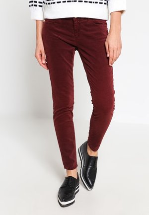 Frau trägt schmal geschnittene burgunderrote Hose, schwarze Plateau-Oxfordschuhe und einen weißen Pullover mit schwarzen Streifen an den Ärmeln.