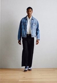 Giacca in denim con lavaggio azzurro chiaro, camicia bianca con colletto, pantaloni neri e scarpe blu lucide. Vestibilità casual con leggere variazioni di texture.