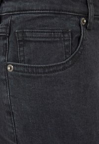 Jeans en denim noir avec une poche arrière ornée d'un clou en métal circulaire. Le tissu présente une texture lisse et un motif de tissage subtil.