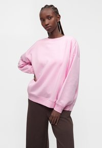 Femme avec des cheveux tressés portant un sweat-shirt rose ample et un pantalon marron, debout avec une main sur la hanche sur un fond uni.
