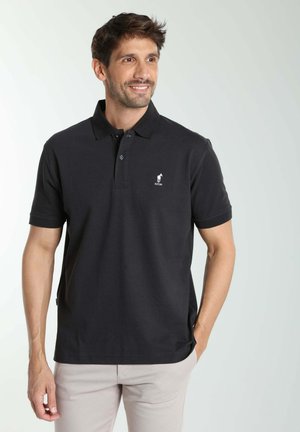 Homme souriant, portant un polo noir à manches courtes avec un petit logo blanc sur la poitrine gauche, et un pantalon de couleur claire, debout avec une main dans la poche.