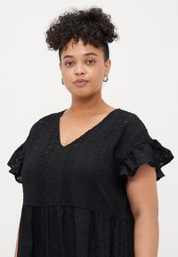 Black cotton dress with a V-neck, short ruffled sleeves, and decorative eyelet patterns throughout the fabric, featuring a flowy silhouette.