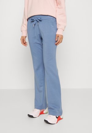 Personne portant un pantalon bleu clair à cordon, un haut à manches longues rose pâle, et des chaussures de course blanches avec des accents rouges et noirs, debout devant un fond uni.