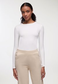Body a maniche lunghe bianco abbinato a pantaloni beige con vita elastica; tessuto morbido con una silhouette aderente. Design semplice, senza motivi visibili.
