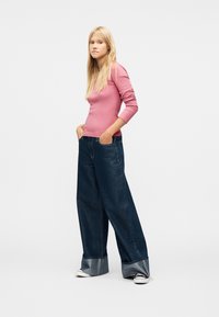 Γυναίκα με μακριά ξανθά μαλλιά που φοράει εφαρμοστό ροζ τοπ μακρυμάνικο, σκούρο τζιν με φαρδιά μπατζάκια και μεγάλες μανσέτες, και ασπρόμαυρα αθλητικά παπούτσια, με τα χέρια στις τσέπες.