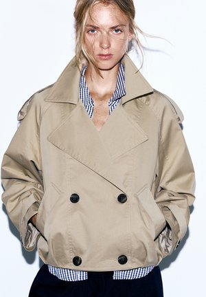 Kvinde med lyst hår iført en beige dobbeltradet trenchcoat over en blå- og hvide ternet skjorte, hænderne i lommerne, foran en ensfarvet hvid baggrund.