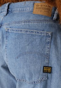 Svjetloplave traperice s kožnom etiketom na kojoj piše "G-Star Originals RAW Denim." Imaju zakrivljeni dizajn stražnjeg džepa.