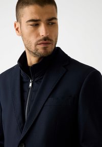 Marineblazer met notch-lapels, gedragen over een zwarte gewatteerde jas. Beschikt over een ritssluiting, een voorzak en een gestructureerde stof.