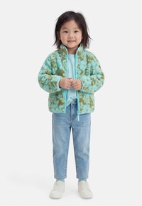 Młode dziecko stojące z złączonymi rękami, ubrane w niebiesko-zieloną polarową kurtkę w wzory, jasnoniebieską koszulę, dżinsy i białe trampki.