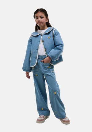 Fille portant une veste bleue et un pantalon avec une broderie de fleurs jaunes, une chemise blanche et des baskets roses et blanches, debout avec la main dans la poche.