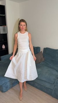 Femme debout en haut sans manches blanc et jupe midi blanche tenant les côtés de la jupe, près d’un canapé bleu et d’une planche de surf à l’intérieur.