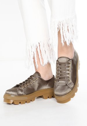 Bronzové saténové tenisky s masivními hnědými gumovými podrážkami. Mají kulaté tkaničky a zlaté očka, spárované s ohrnutými bílými kalhotami po kotníky.