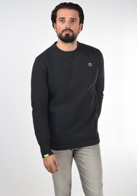 Black sweatshirt with a crew neckline, textured fabric, and a small green logo on the chest. Features ribbed cuffs and hem. Paired with light grey jeans.