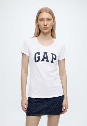 Жена със прави блондини коси, носеща бяла тениска GAP и тъмносиня дънкова пола с диамантен мотив, стояща на едноцветен фон.