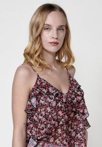 Camisola floreale in tessuto nero trasparente, con bordi arricciati e fiori rosa. Spalline sottili e una vestibilità ampia enfatizzano il suo design leggero.
