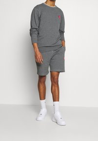 Muž v šedé mikině s dlouhým rukávem a odpovídajících šortkách s červenými logy, bílé ponožky a bílé tenisky, stojící před jednobarevným pozadím.